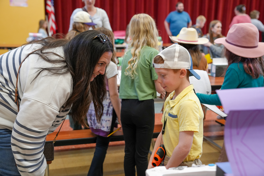 An adult leans down to speak with a young student wearing a hat and holding a clipboard during a classroom project display, with other students and families in the background.