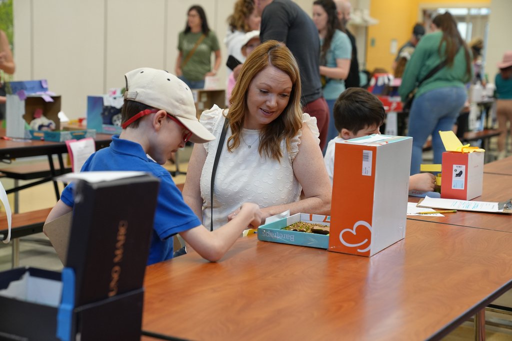 An adult sits beside a student at a table, looking at a shoebox habitat project together while other displays and participants fill the room behind them.