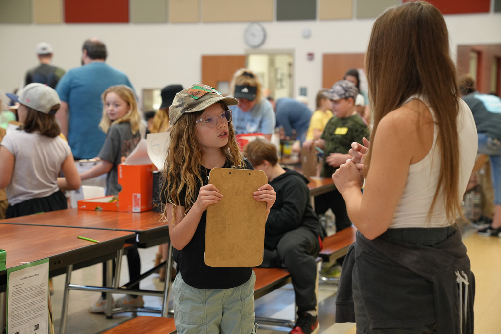 A student wearing a hat holds a clipboard while speaking with an adult in a school cafeteria, as other students and families present projects in the background.