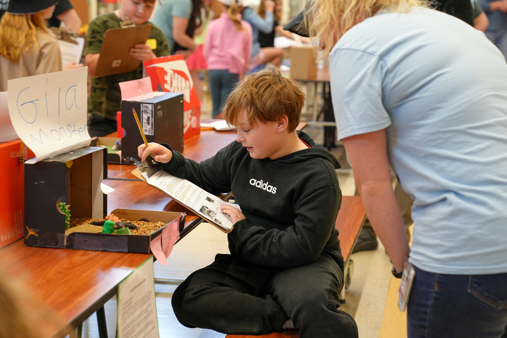 A student sits at a cafeteria table writing on a clipboard while presenting a shoebox habitat project, as an adult leans in to observe.
