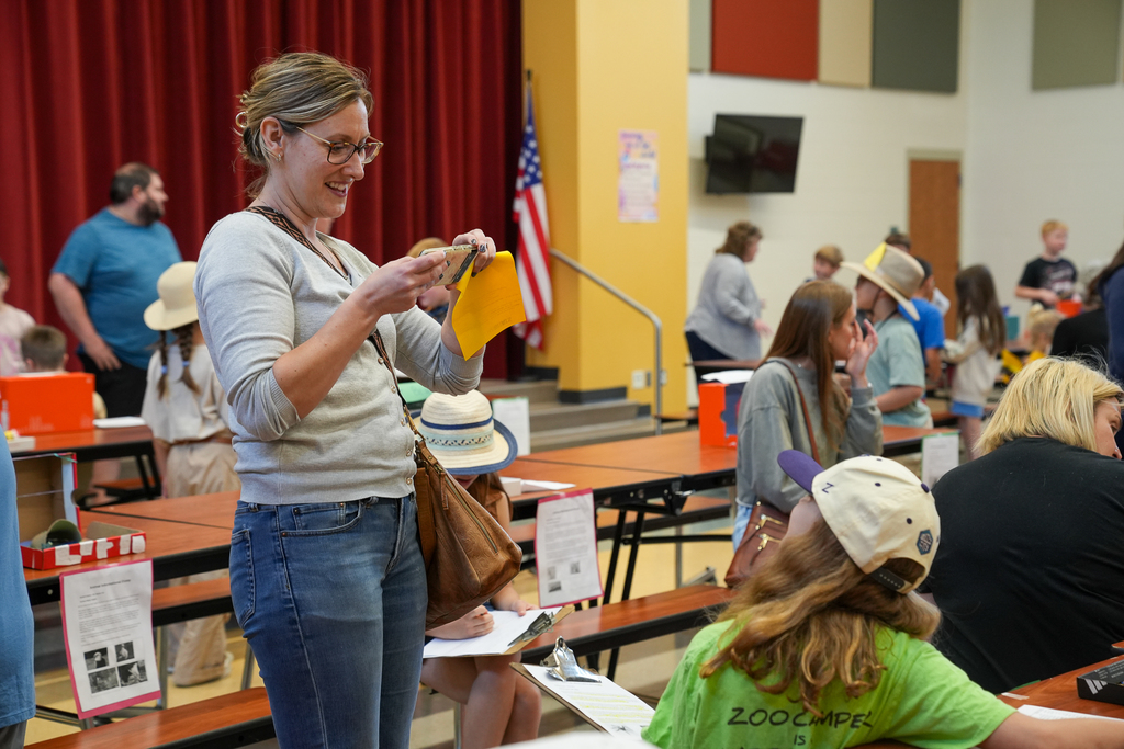 An adult stands in a school cafeteria taking a photo of student projects displayed on tables, while families and students explore exhibits around the room.