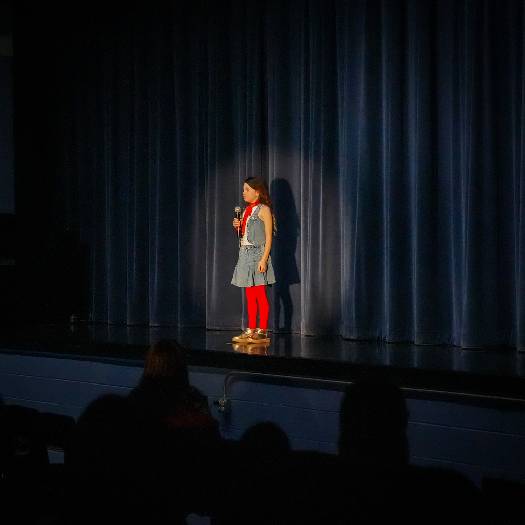 A student stands on stage holding a microphone, wearing a denim outfit with bright red tights, preparing to perform in front of an audience.