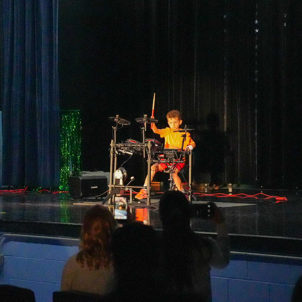A young student plays an electronic drum set on stage, holding drumsticks mid-performance while audience members watch and record from their seats.