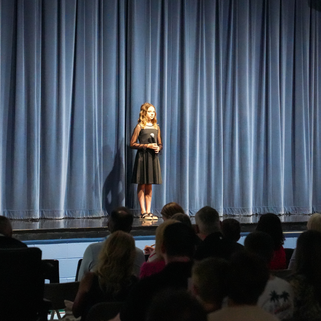 A student stands center stage holding a microphone, wearing a black dress, preparing to perform in front of a full audience.