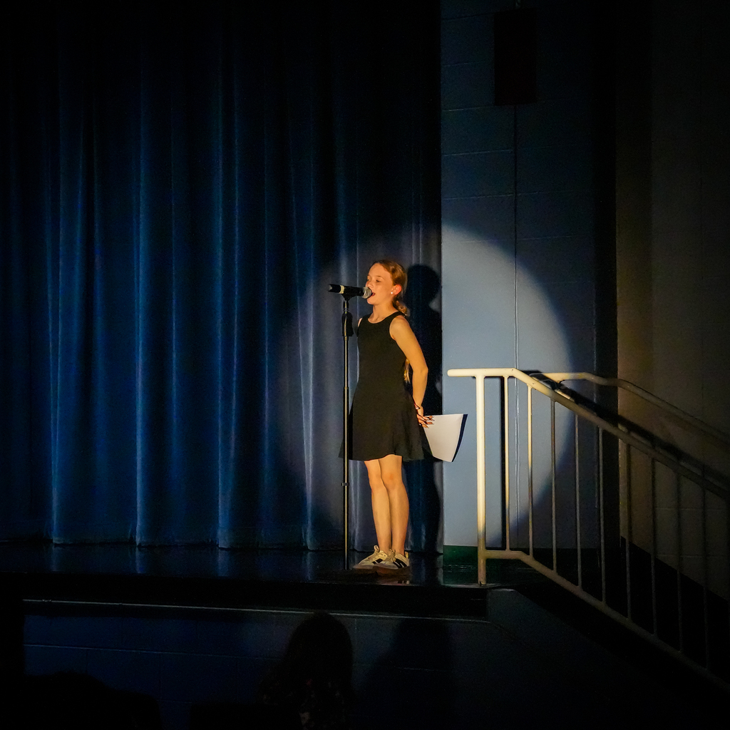 A student stands on stage singing into a microphone under a spotlight, holding a paper in one hand during a talent show performance.