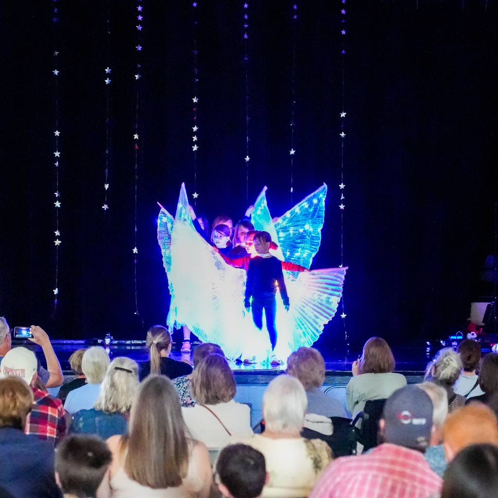 A group of students performs on stage using glowing light props, creating bright, wing-like shapes in front of a seated audience at a school talent show.