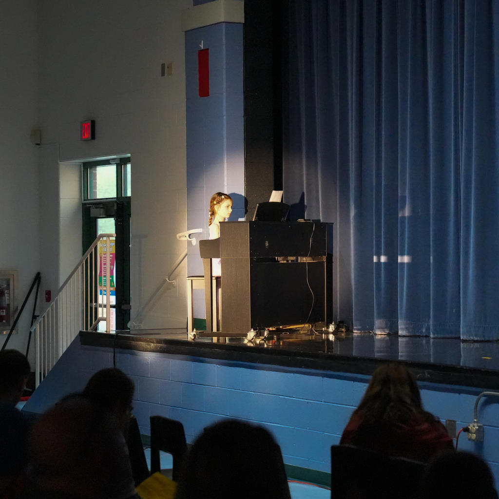 A student sits at a piano on stage, focused on playing while a spotlight shines on them and audience members watch quietly.