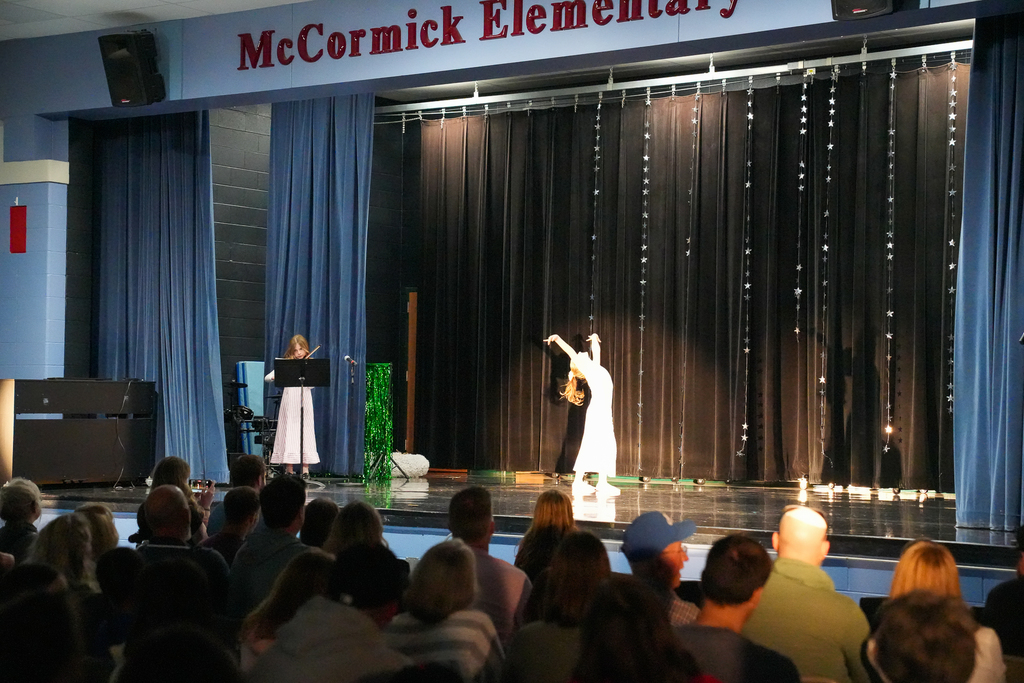 A student dances on stage under bright lights during McCormick Elementary’s talent show while another student plays an instrument in the background, with an audience watching from their seats.