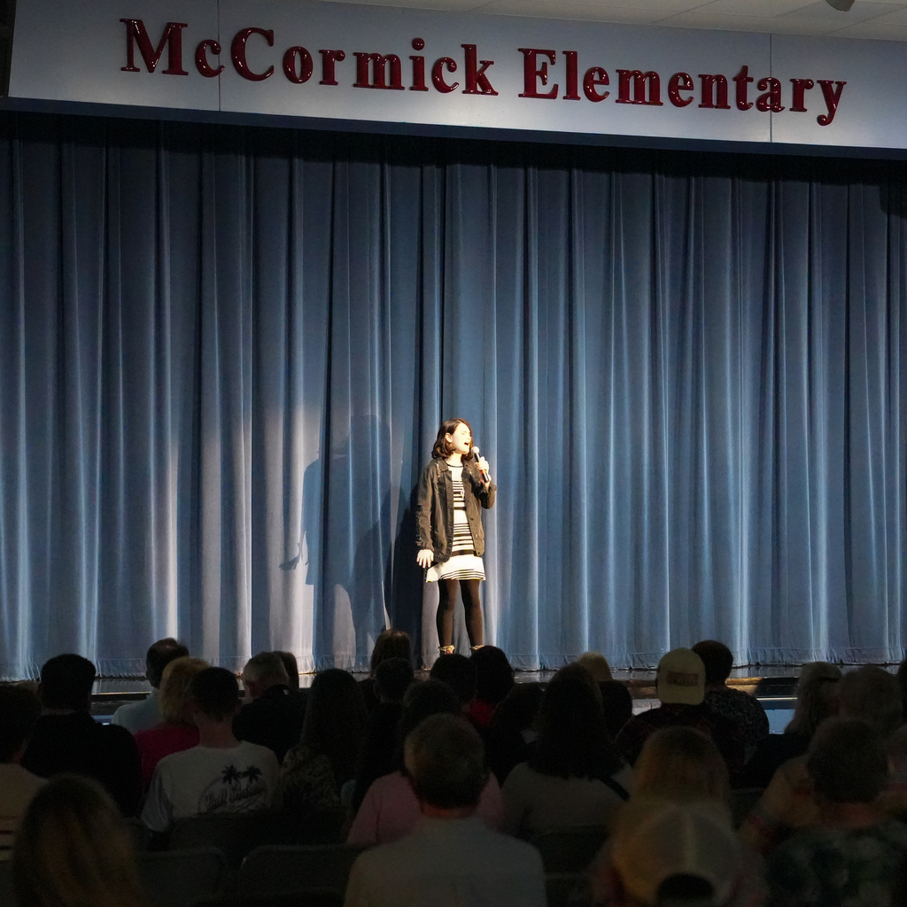 A student stands on stage under a spotlight speaking or singing into a microphone, with a curtain backdrop and audience seated below.