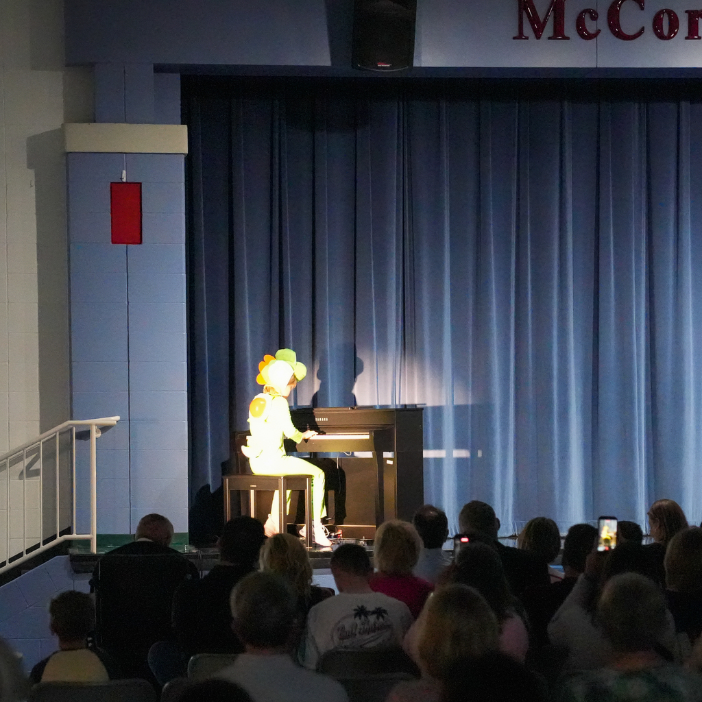 A student in a Yoshi costume sits at a piano on stage, performing under a spotlight while the audience watches.