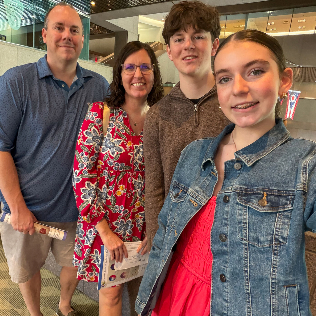 A group of four people, including two adults and two students, smile for a selfie inside a building, with one person holding a program.