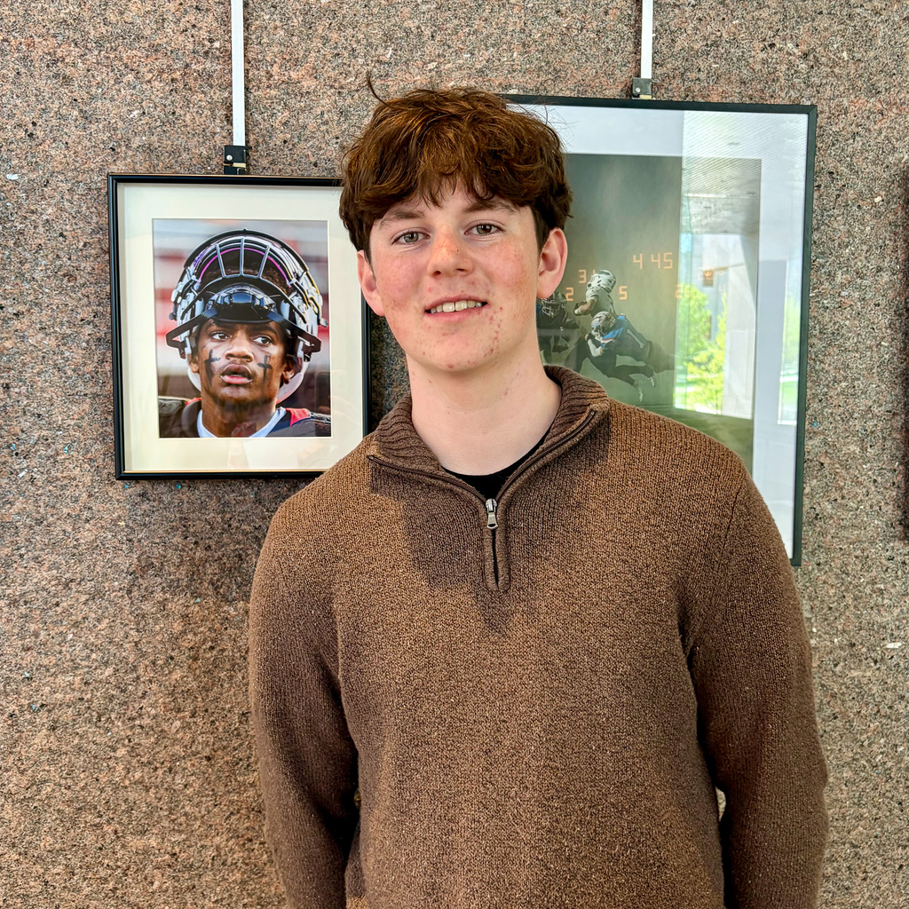 A high school student with brown hair wearing a brown quarter-zip sweater stands in front of a granite wall displaying framed sports photography.