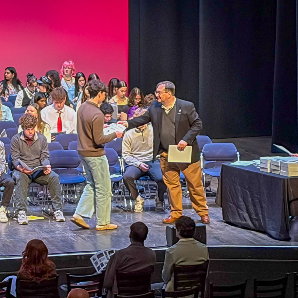 A student walks across a stage to receive a certificate from an adult during an award ceremony, while other students sit behind them and audience members watch from the front rows.