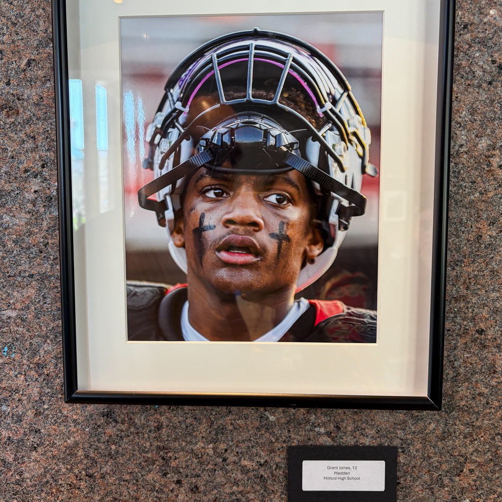 A framed portrait of a football player wearing a helmet and eye black with cross markings on his cheeks is displayed on a wall, with a nameplate below reading “Grant Jones, 12, Madden, Milford High School.”