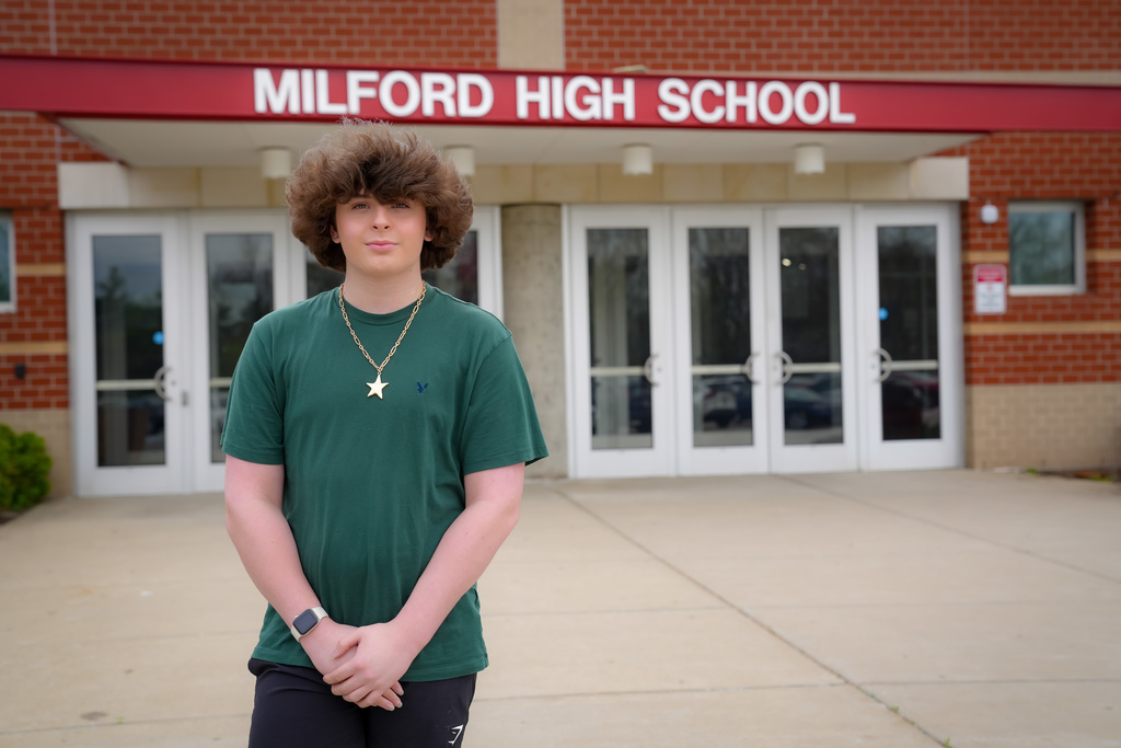 A student stands outside the entrance of Milford High School, with the school name displayed above the doors in red lettering. The student is wearing a green shirt and a necklace, standing with hands clasped in front, facing the camera. The background shows the school’s brick exterior and glass entry doors.
