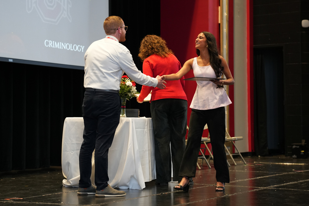 A student shakes hands with a staff member while holding an award during the ceremony. A presentation slide behind them references a field of study in criminology.
