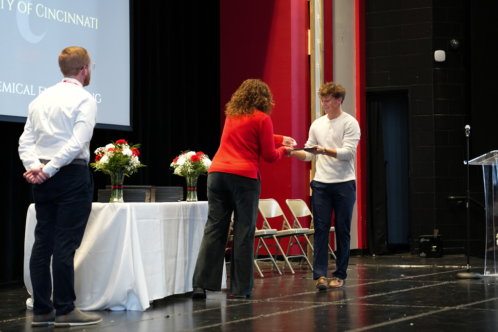 A student accepts an award on stage from a staff member while another stands nearby. A screen behind them references the University of Cincinnati and a field of study in chemical engineering.