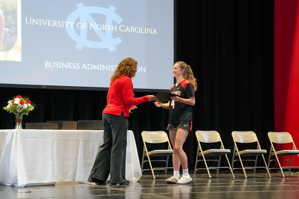A staff member presents an award to a student on stage during the ceremony. The student smiles while receiving the certificate as a screen behind them displays “University of North Carolina” and “Business Administration.”