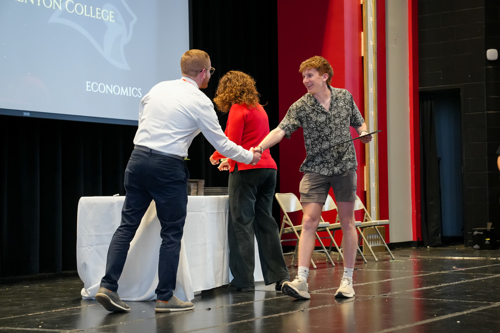 A student smiles while shaking hands with a staff member after receiving an award. A presentation slide behind them references Kenyon College and economics.