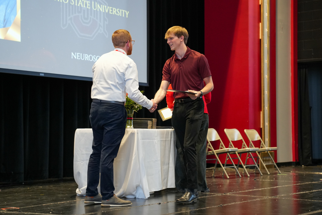 A student shakes hands with a staff member while holding an award. A screen behind them references The Ohio State University and neuroscience.