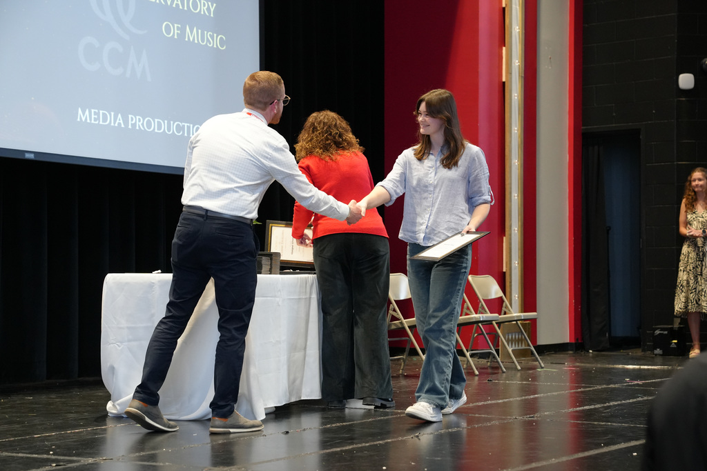 A student smiles while shaking hands with a staff member after receiving an award. A presentation slide behind them references the Cincinnati Conservatory of Music and media production.