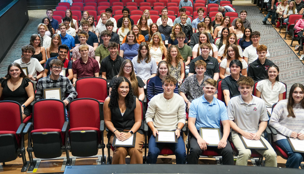 Group photo of Milford High School Class of 2026 Summa Cum Laude students seated in rows inside an auditorium. Students are dressed in semi-formal attire, smiling toward the camera, with several in the front row holding framed awards.