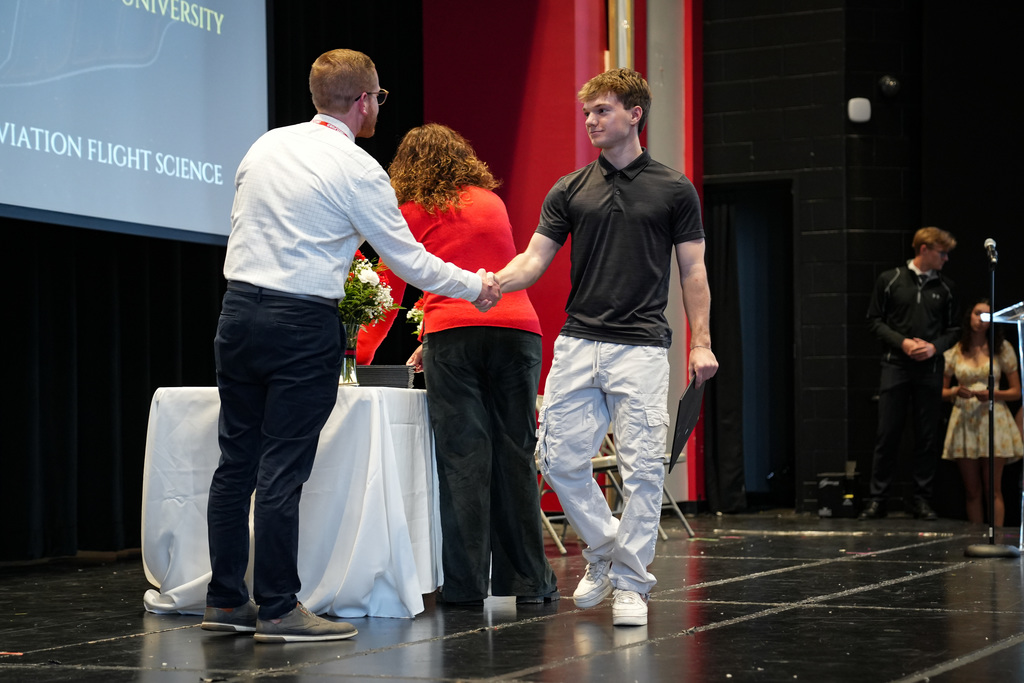 A student receives an award and shakes hands with a staff member on stage during the ceremony. The screen behind them references aviation flight science.