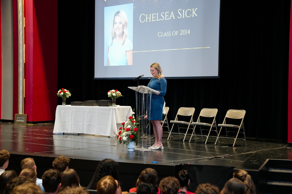 A woman stands at a clear podium on a stage addressing an audience during Milford High School’s Summa Cum Laude Academic Convocation. A large screen behind her displays her name, “Chelsea Sick, Class of 2014.” A table with stacked awards and floral arrangements sits nearby.