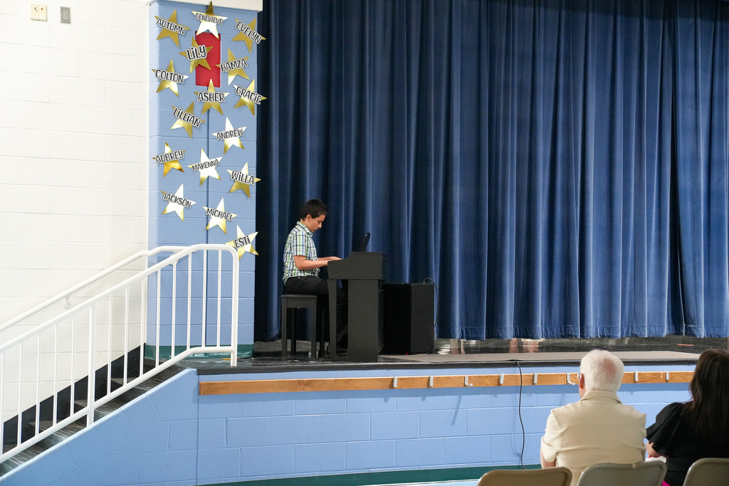 Student plays a keyboard on stage during a talent show with a blue curtain backdrop and audience seated in front.