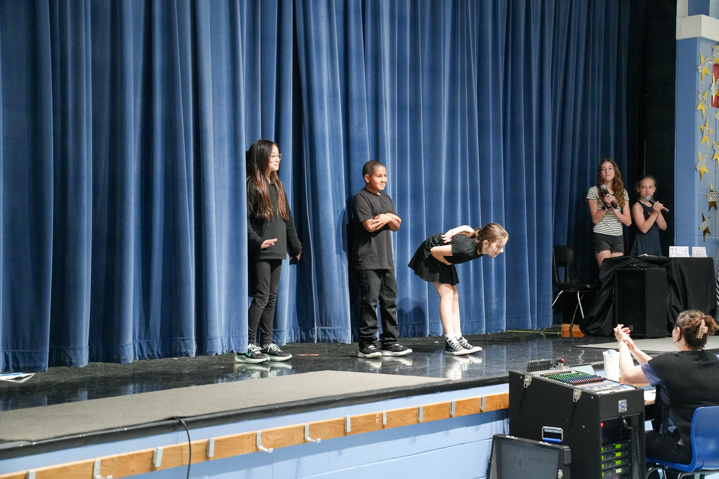 Several students stand on stage taking a bow at the end of their performance while others hold microphones to the side.