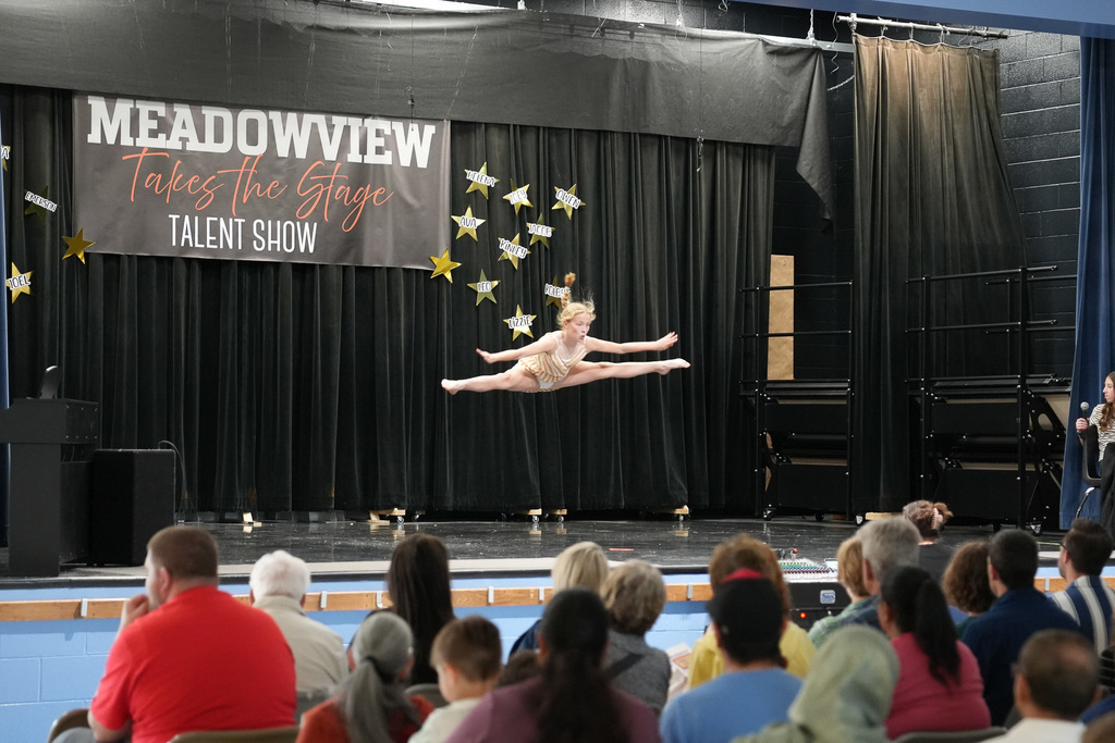 Student performs a high leap across the stage during a dance routine at the Meadowview talent show while the audience watches.