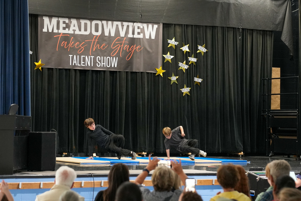 Two students in black outfits perform a coordinated floor routine on stage while audience members watch and take photos.