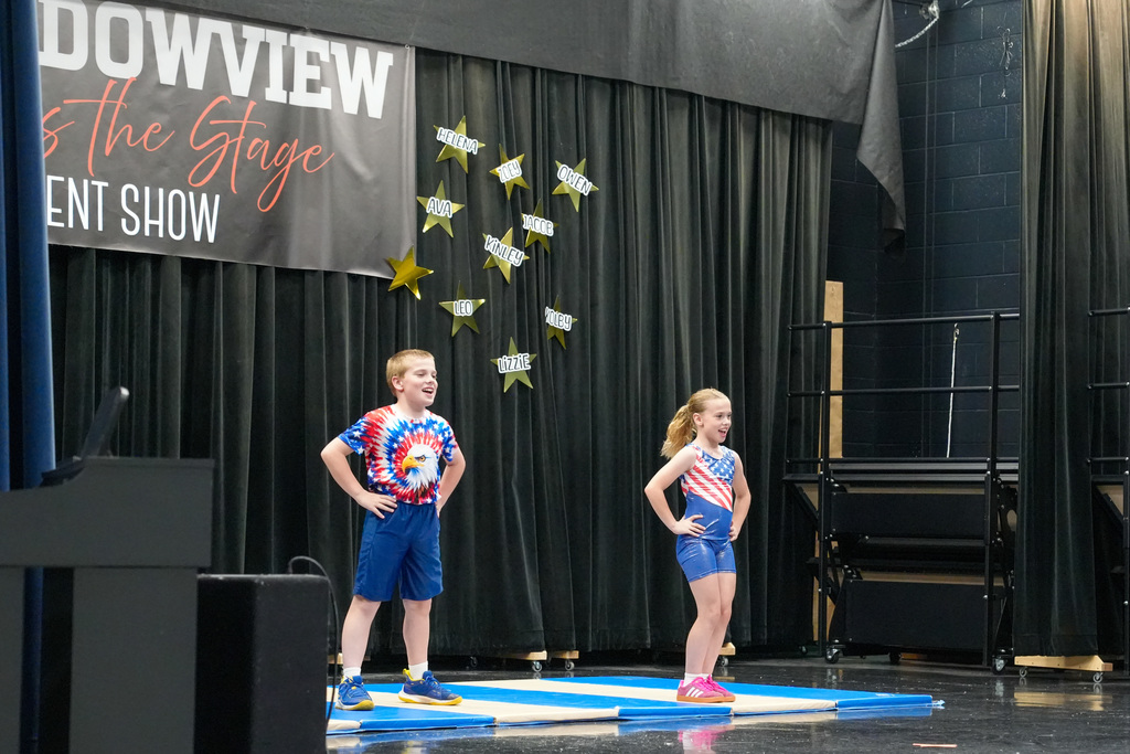 Two elementary students perform a gymnastics or dance routine on mats on stage beneath a “Meadowview Takes the Stage Talent Show” banner.