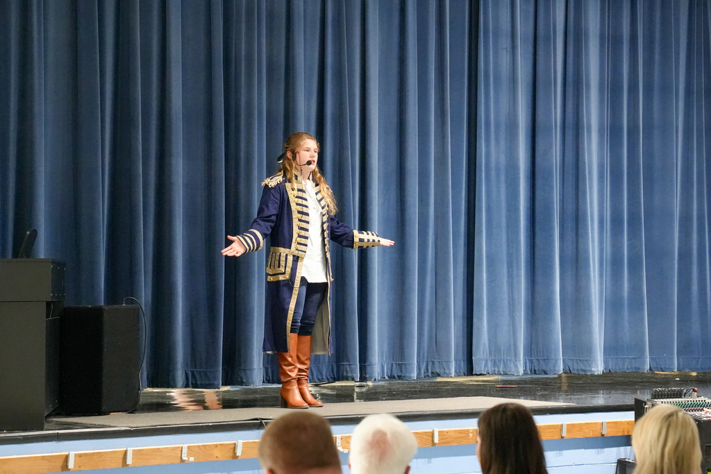 Student in a historical-style costume with a headset microphone performs on stage in front of a blue curtain during a school talent show.