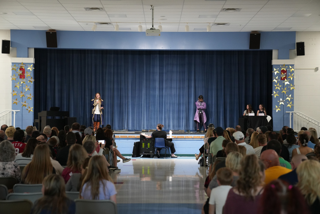 Wide view of a school cafeteria stage during a talent show with two student performers in costume, a sound technician at a control board, and a full audience seated in rows.