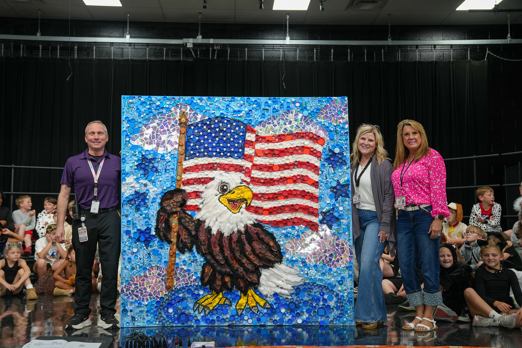 Three adults stand beside a large collaborative student mural featuring a bald eagle in front of an American flag. The artwork is made from colorful found objects such as bottle caps, beads, and tiles, creating a textured mosaic effect. Students sit in the background on a stage, watching the presentation.