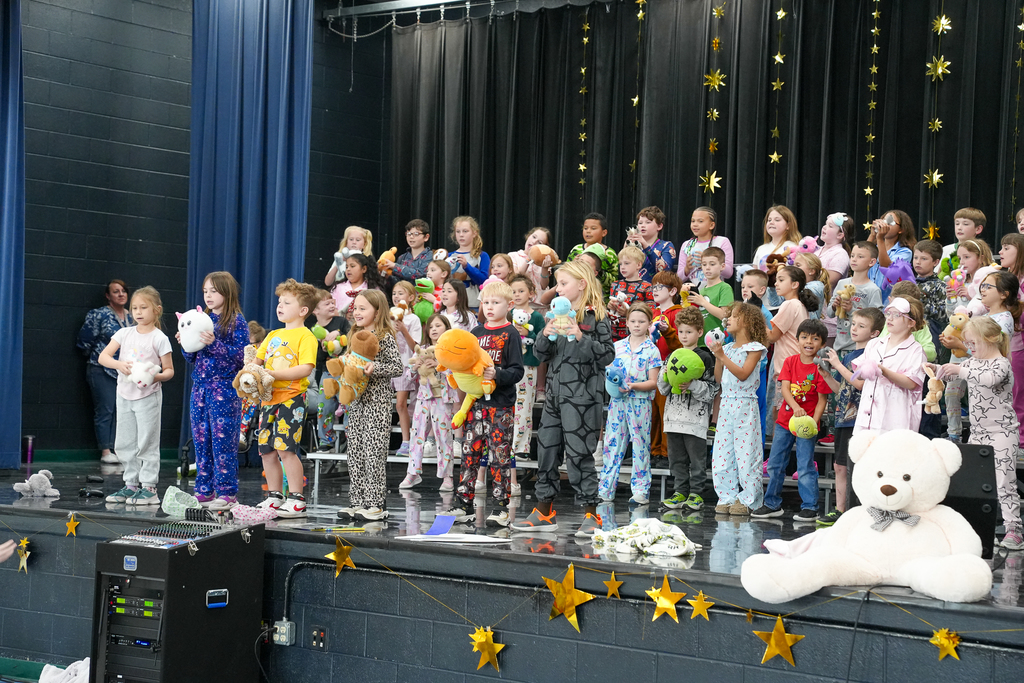 A large group of elementary students in pajamas sing together on risers, holding stuffed animals as part of a themed school performance.