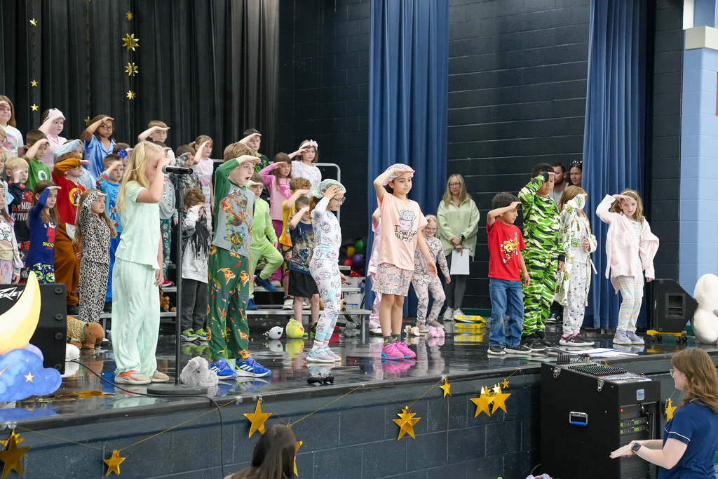 Students in pajamas perform synchronized movements on stage under hanging star decorations during a school program.