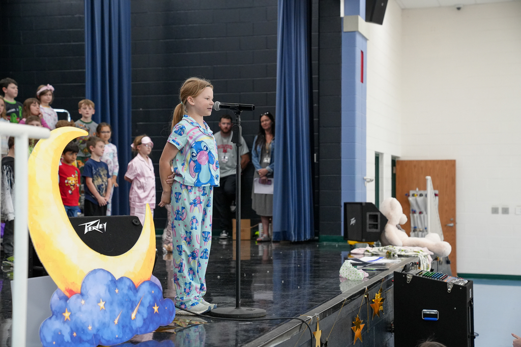 A student in colorful pajamas stands at a microphone on stage, performing in front of classmates and staff during a school musical.