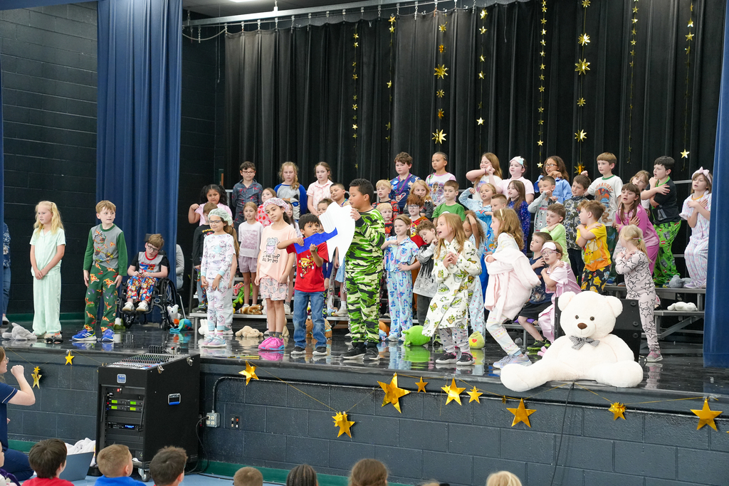 A large group of elementary students in pajamas sing together on risers, holding stuffed animals as part of a themed school performance.