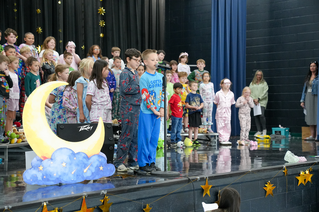 Elementary students in pajamas stand on risers on a school stage decorated with stars and a crescent moon, while two students speak into a microphone during a performance.