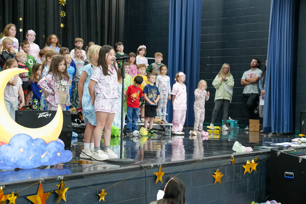 A young student in pajamas sings into a microphone at center stage, with classmates in pajamas standing behind her during a school performance.