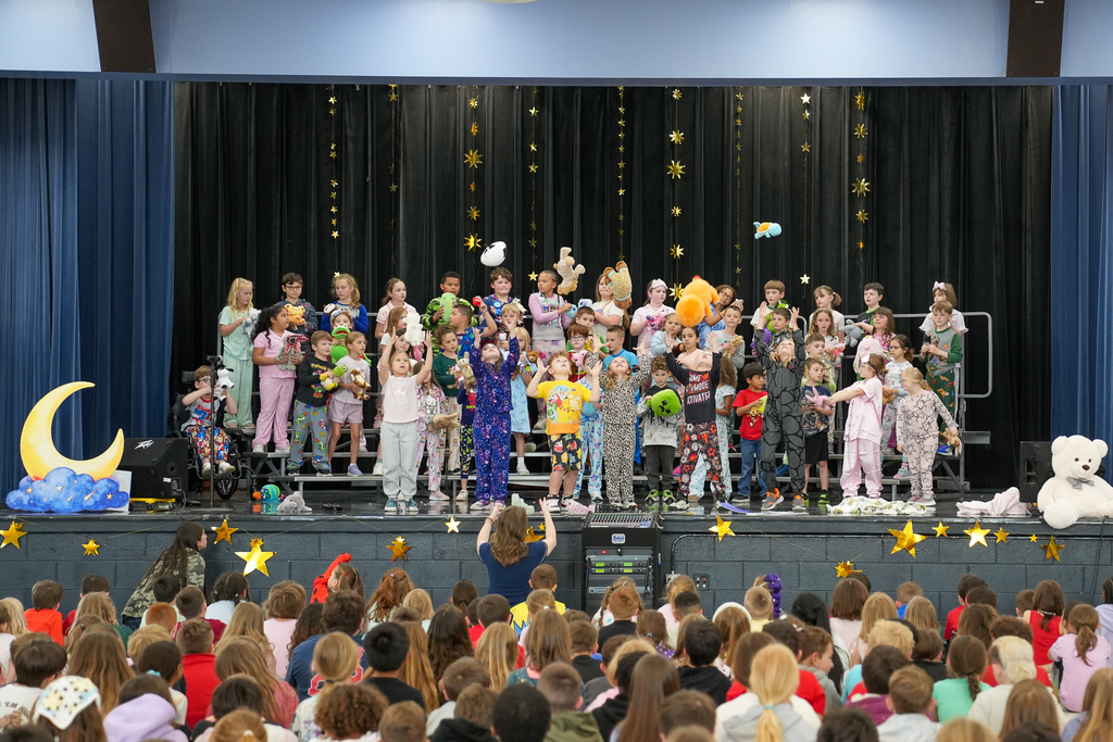 Students toss stuffed animals into the air on stage during a lively pajama-themed school performance, with an audience seated in front.