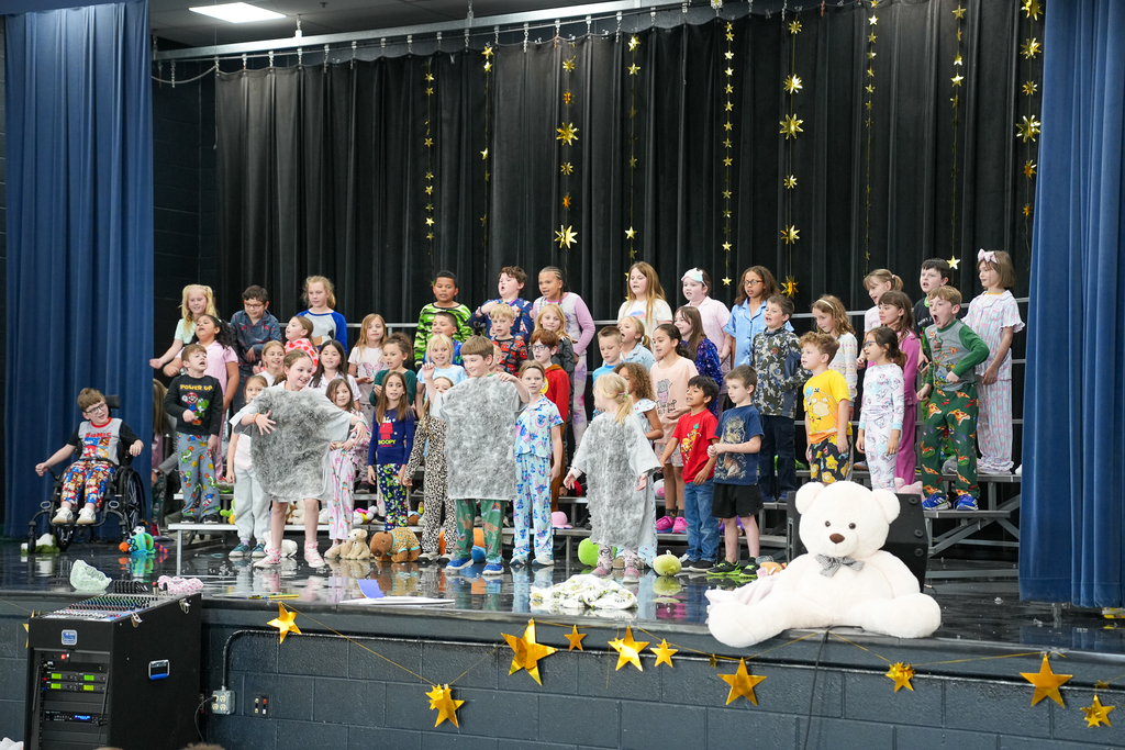 A group of students in pajamas dance in formation on stage, performing for classmates seated on the floor in front of them.