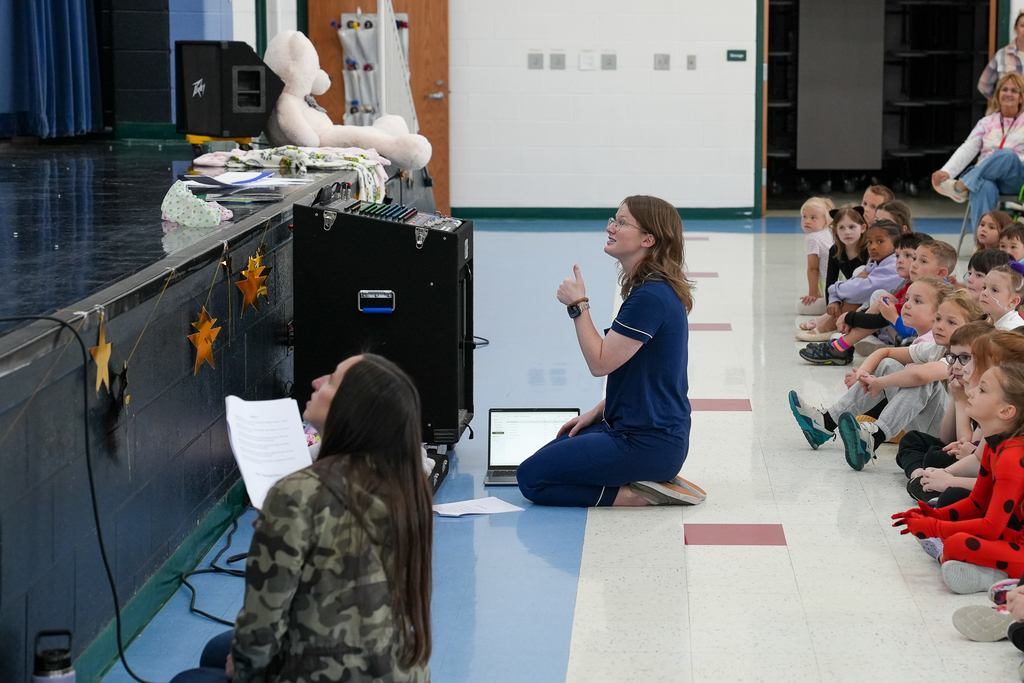 A teacher kneels near the stage giving cues to seated students watching a pajama-themed school performance, with props and sound equipment nearby.