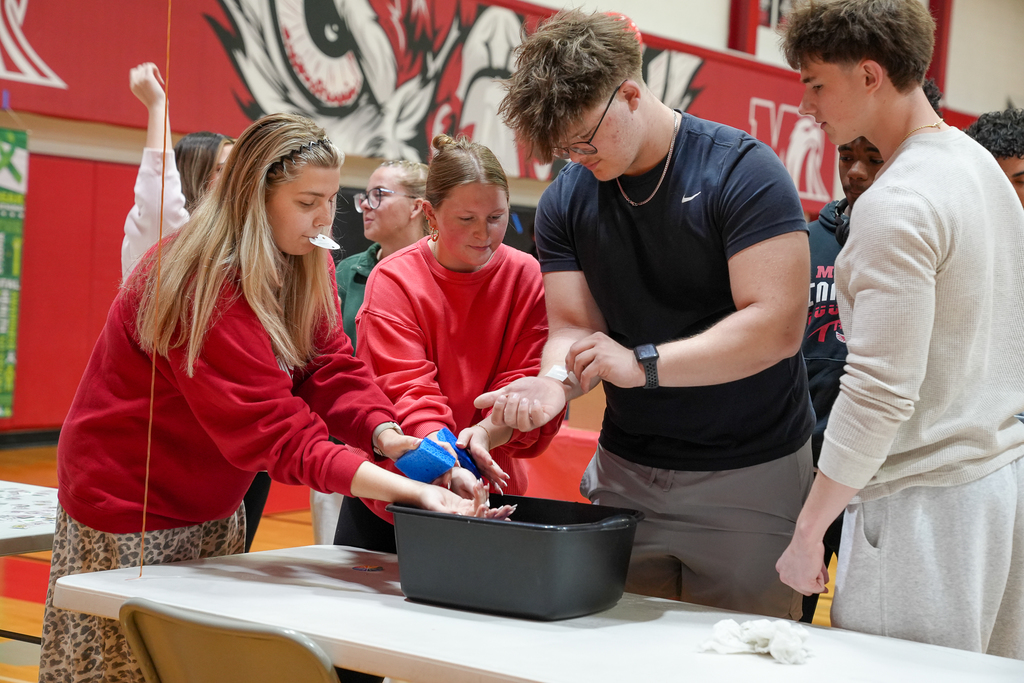 A group of students gather around a table, washing or rinsing their hands in a bin as part of an interactive activity.