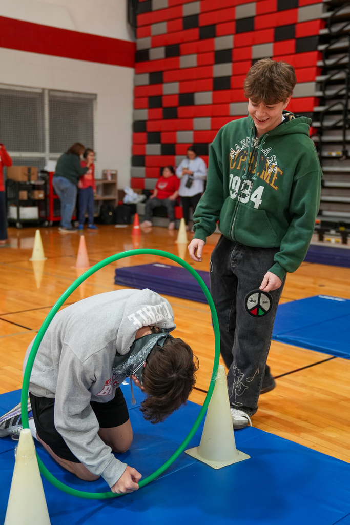 A student kneels on a mat and crawls through a green hula hoop set between cones while another student smiles and watches in a school gym.