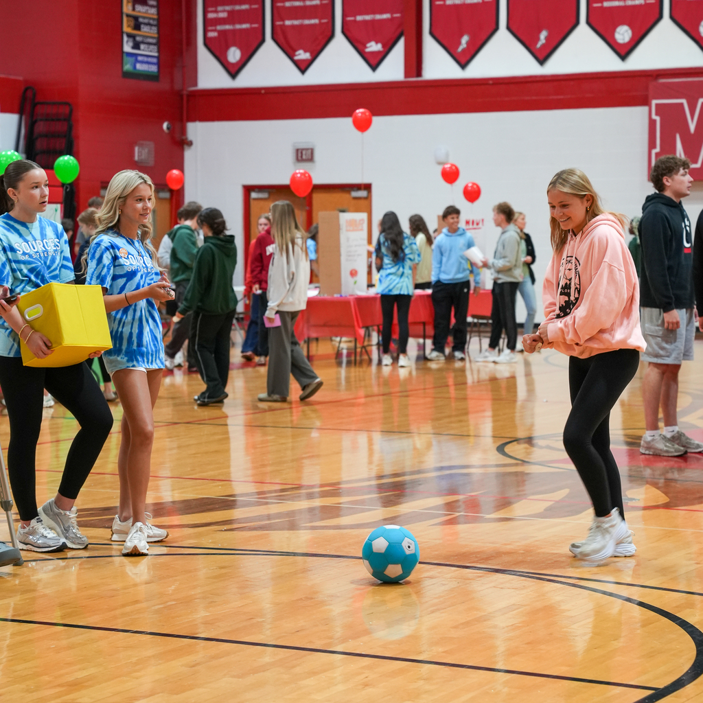 Students play a game in the gym involving a blue ball, with others and activity stations visible in the background.