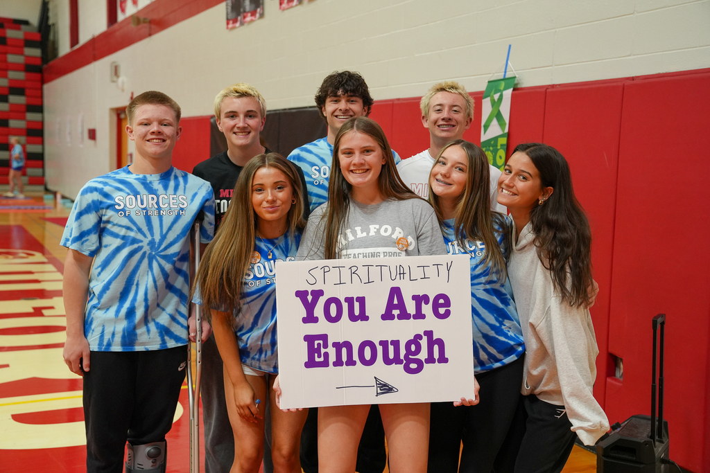 A group of smiling high school students pose together in a gym holding a sign that reads “You Are Enough.”