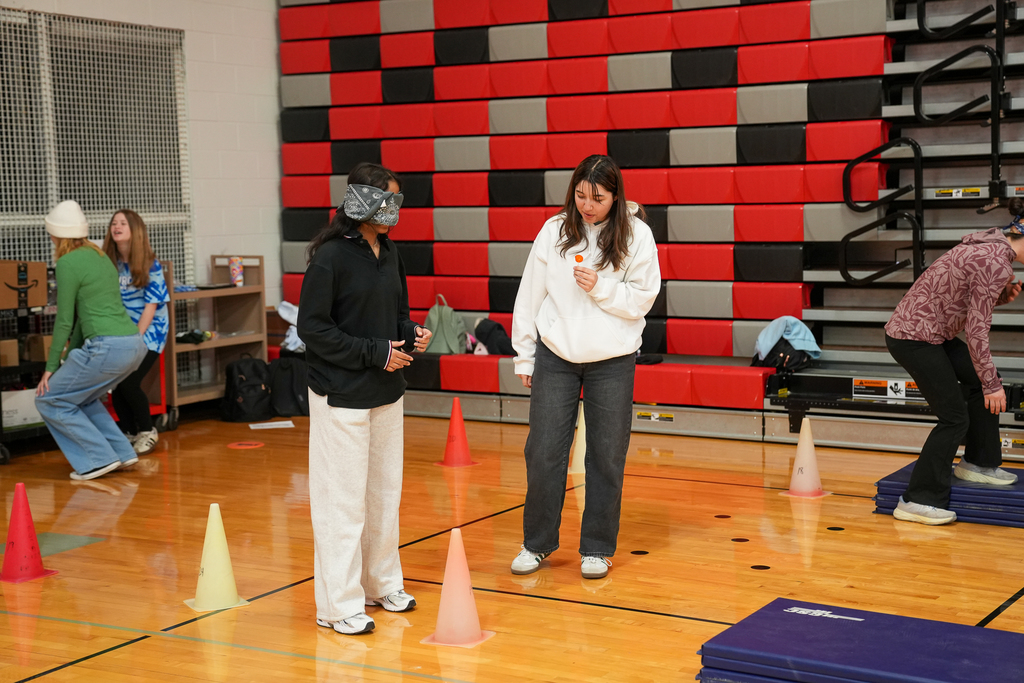 Two students stand near cones on the gym floor, one blindfolded, as they participate in a guided movement challenge.